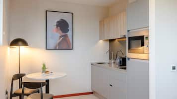 A compact kitchen with a microwave, sink, and cabinets. There's a round table with chairs and a small plant. A framed picture hangs on the wall.