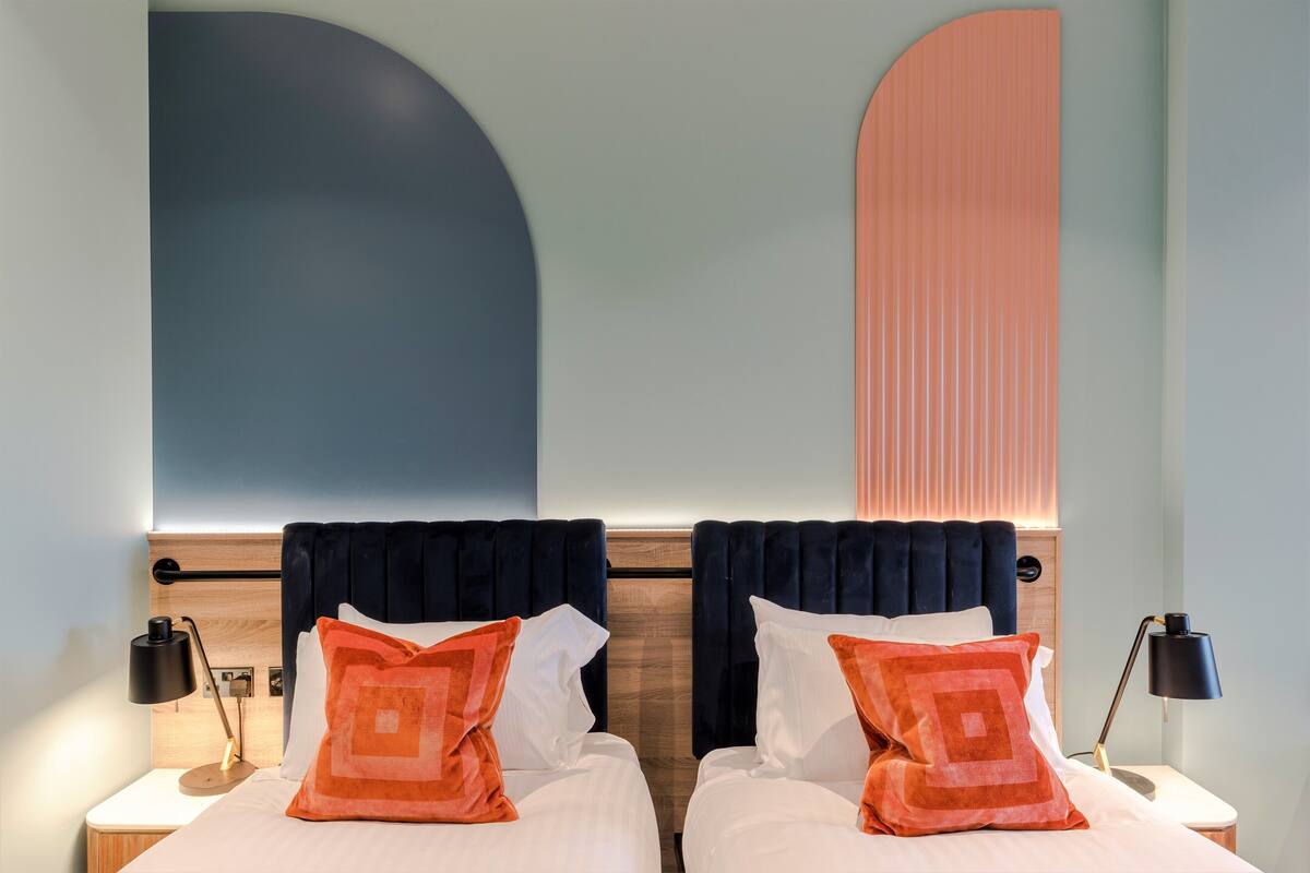 Une chambre d’hôtel avec deux lits, chacun doté d’une tête de lit sombre, de draps blancs et d’oreillers orange. On y trouve des tables de chevet avec des lampes, et une grande décoration murale abstraite au-dessus des lits.