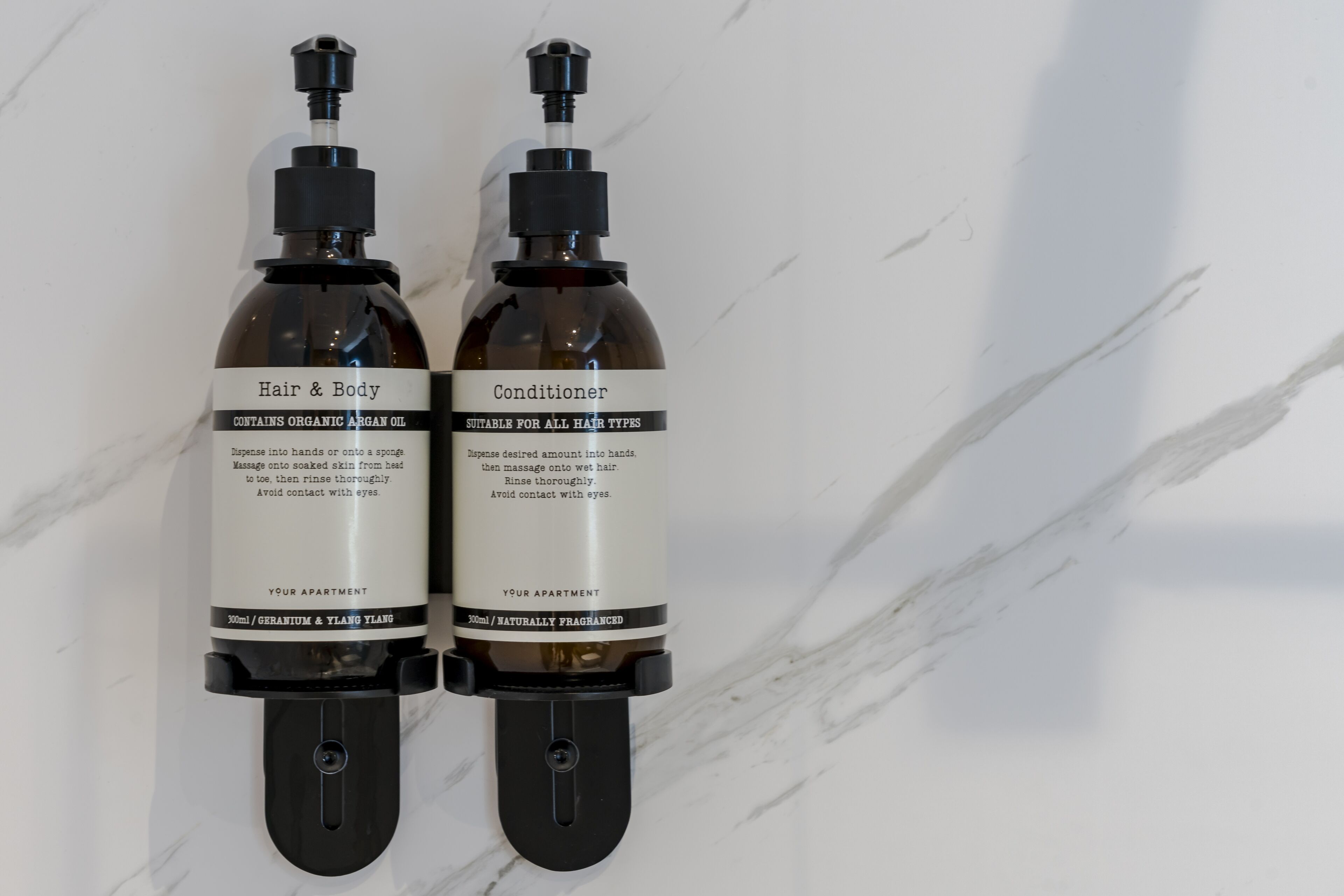 Deux flacons d’après-shampoing et de soin pour le corps, posés sur une surface en marbre.