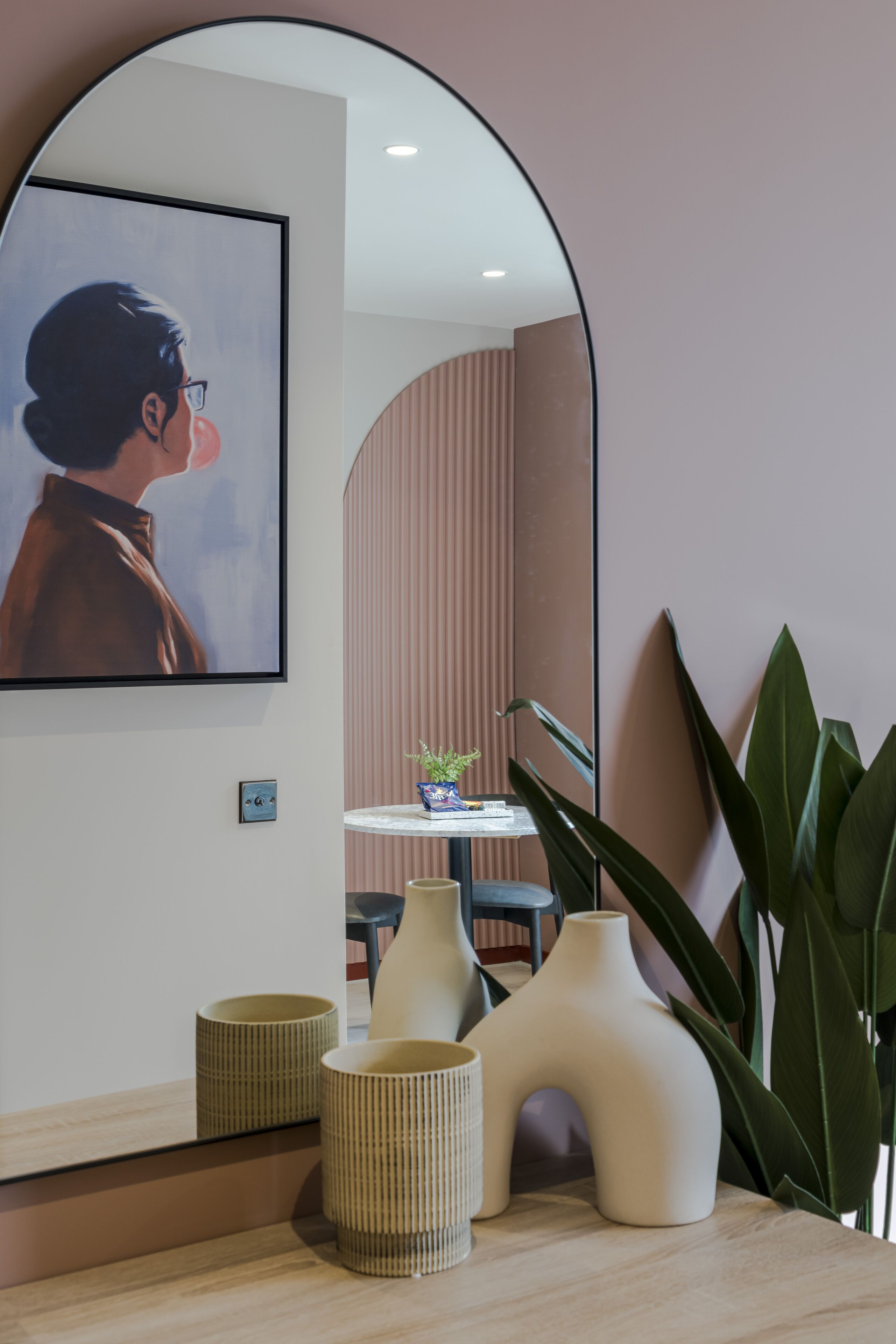 A modern interior with a large mirror reflecting a framed artwork, a small table with a plant, and decorative vases on a wooden surface.