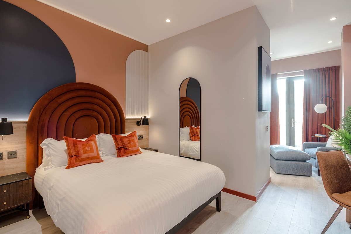 Une chambre d’hôtel moderne dotée d’un grand lit, d’une tête de lit incurvée, d’un miroir et d’un coin salon avec un fauteuil et une petite table.