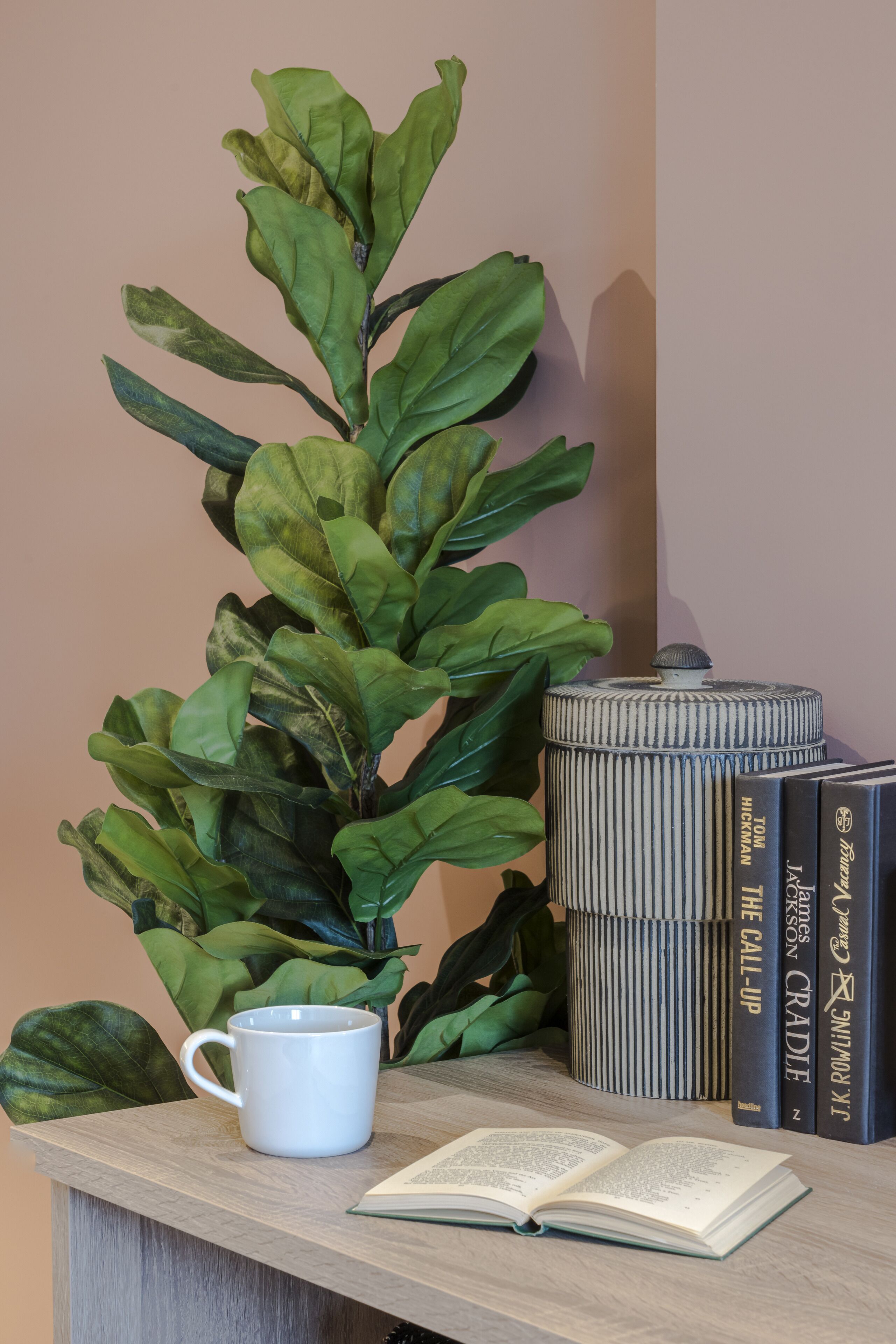 Una mesa con una planta en maceta, una taza blanca, un libro abierto y una pila de libros.
