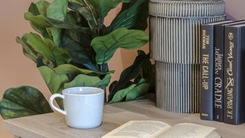 Una mesa con una planta en maceta, una taza blanca, un libro abierto y una pila de libros.