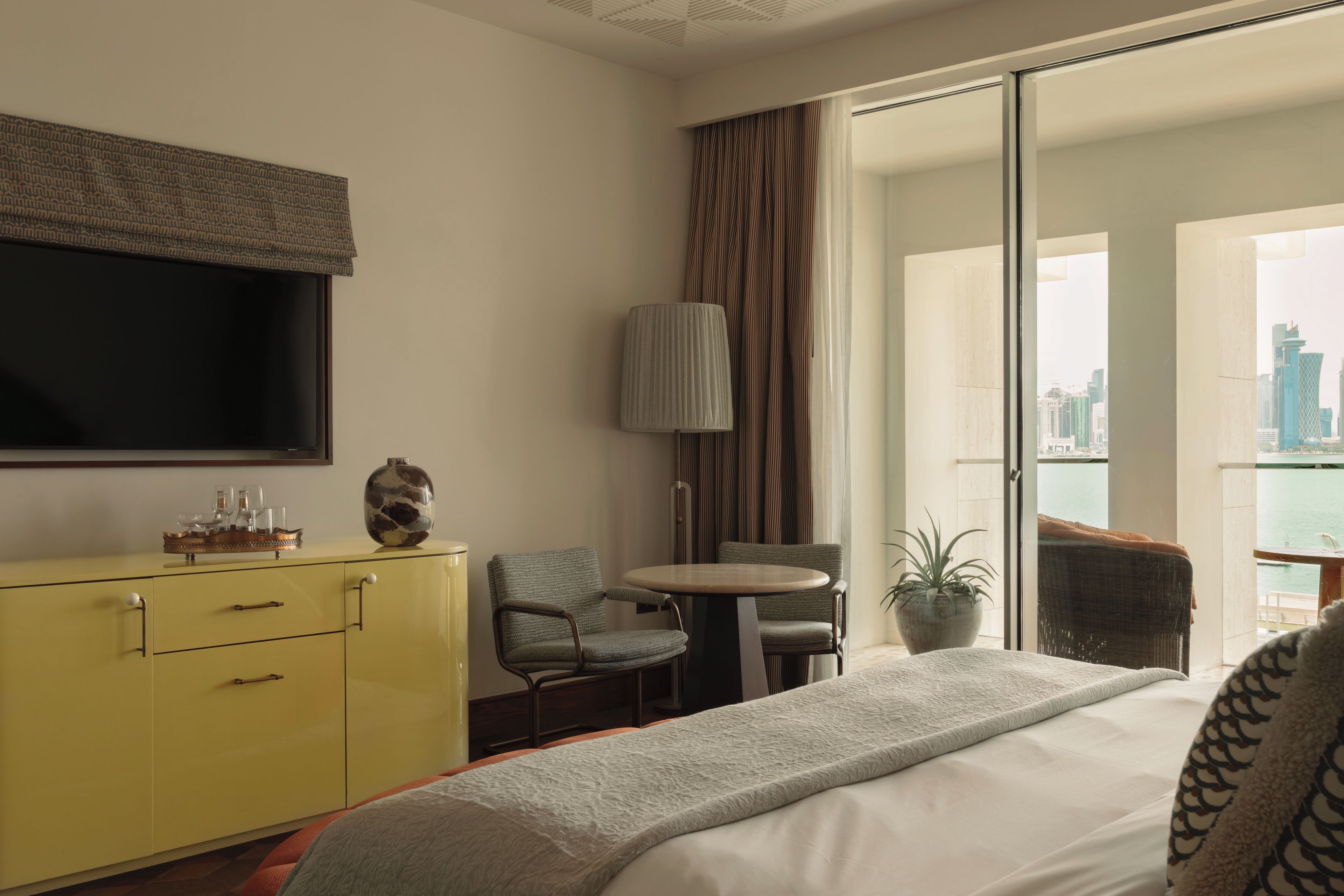 A hotel room with a bed, a yellow dresser, a television, a chair, a table, a lamp, and a view of the city.