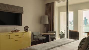 A hotel room with a bed, a yellow dresser, a television, a chair, a table, a lamp, and a view of the city.