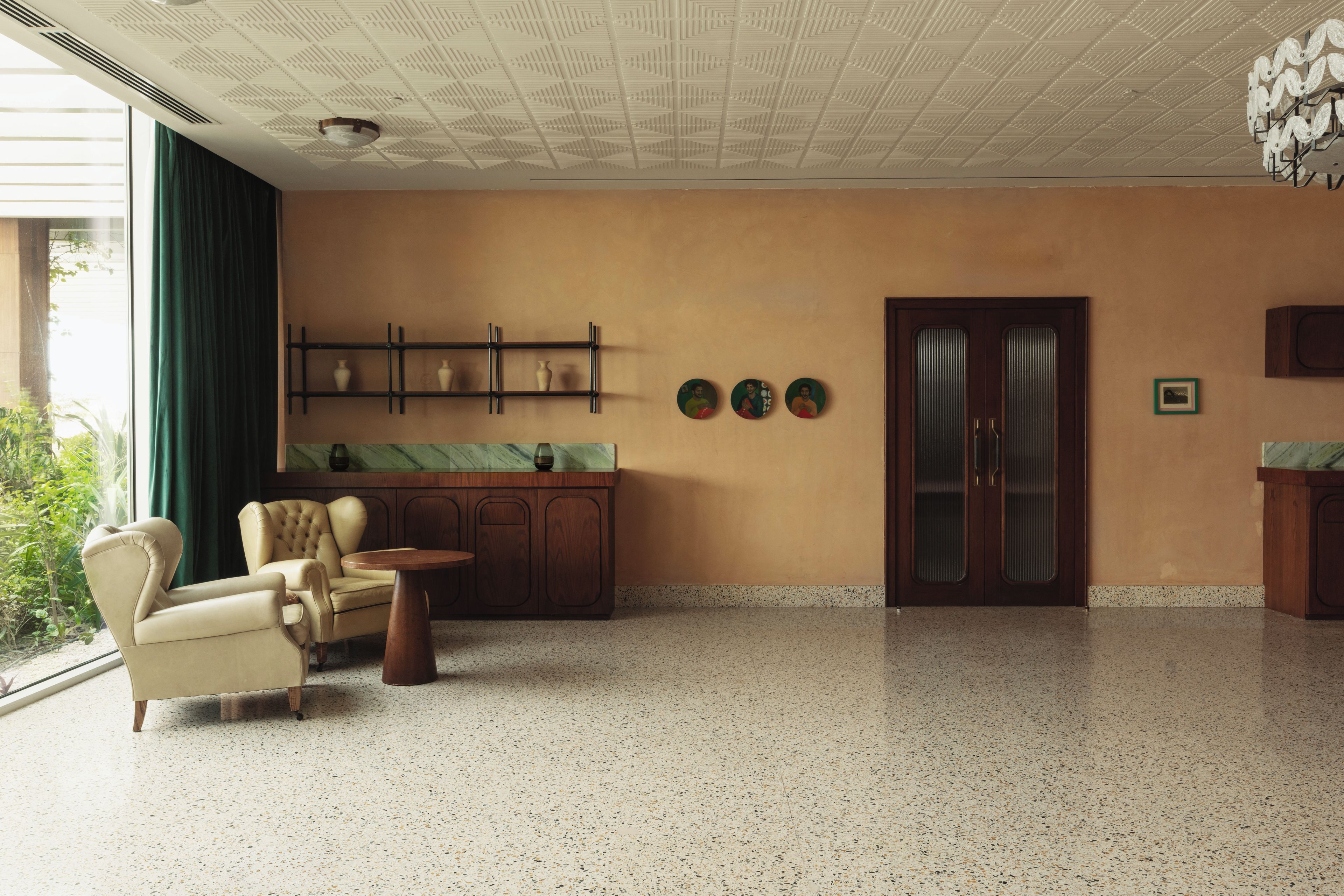 A spacious room with a large window, a seating area with a chair and a small table, a wooden cabinet with decorative items, and a door with a frosted glass panel.