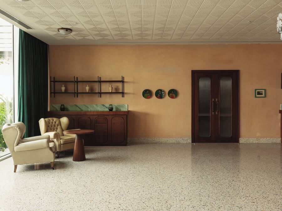 A spacious room with a large window, a seating area with a chair and a small table, a wooden cabinet with decorative items, and a door with a frosted glass panel.