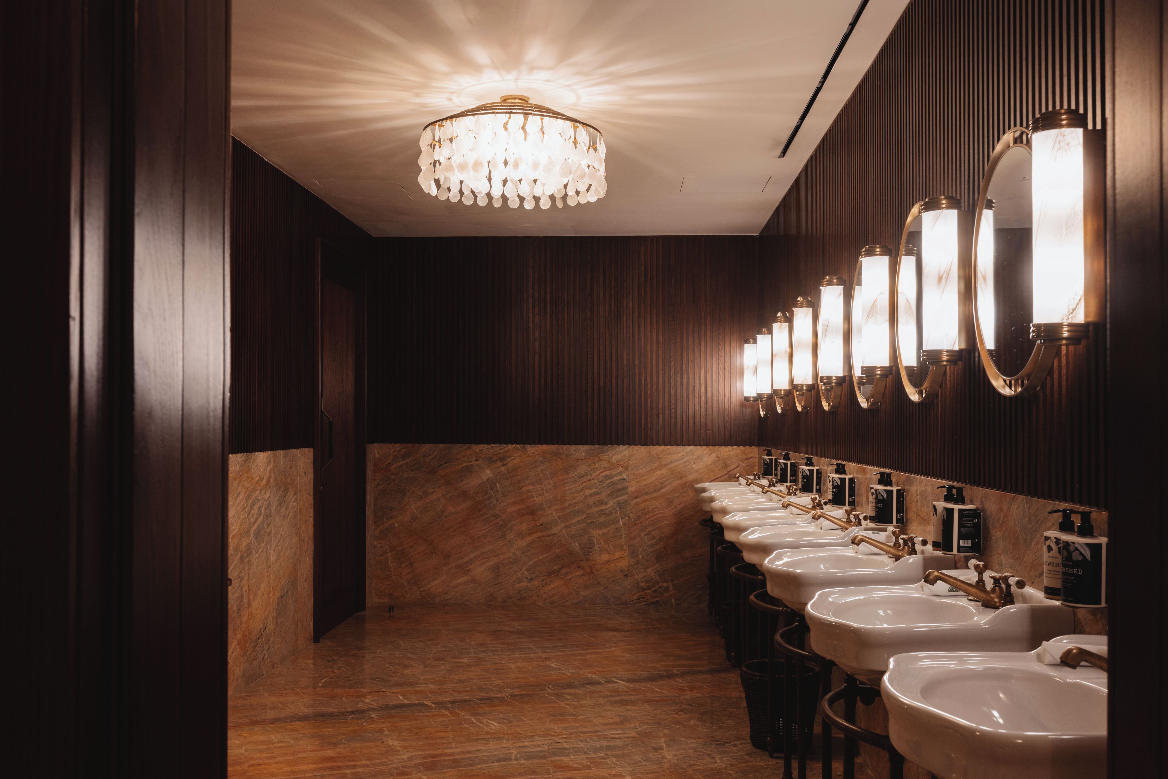 A row of sinks with mirrors and soap dispensers in a restroom with wooden paneling and a marble floor.
