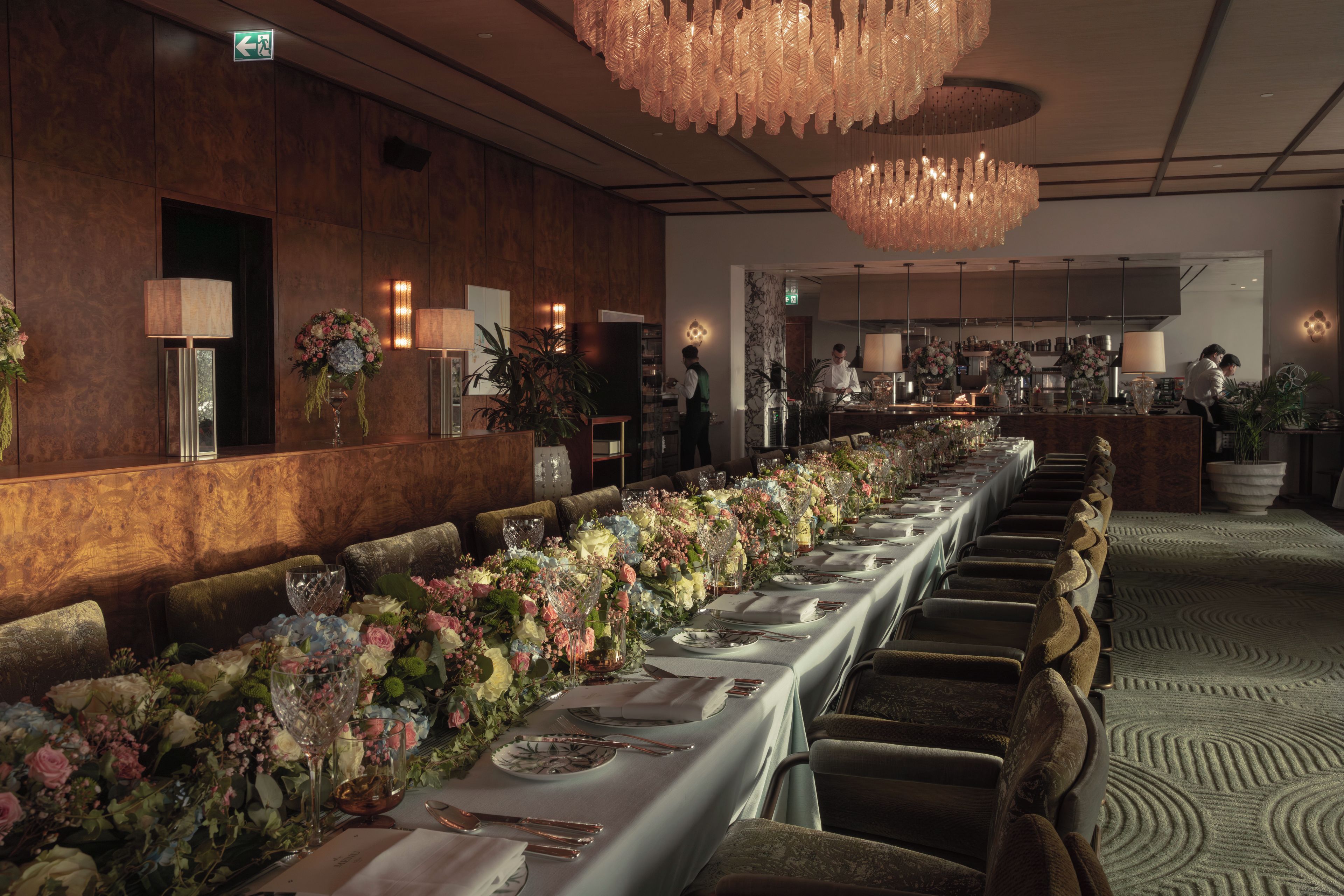 Uma longa mesa de banquete decorada com arranjos florais, louças elegantes e um lustre moderno.
