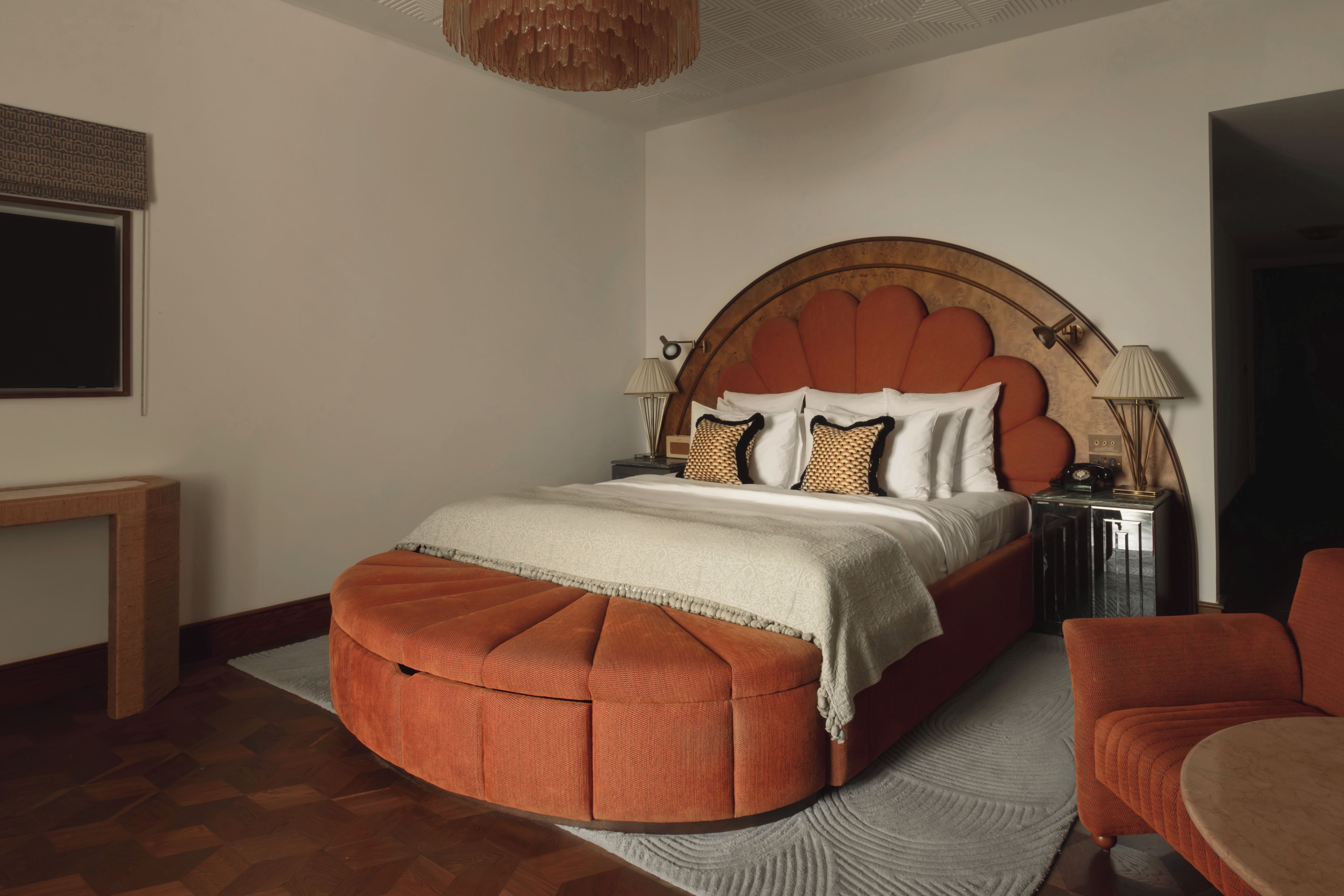 A bedroom with a large bed, a bedside table with lamps, a chair, and a wooden floor.