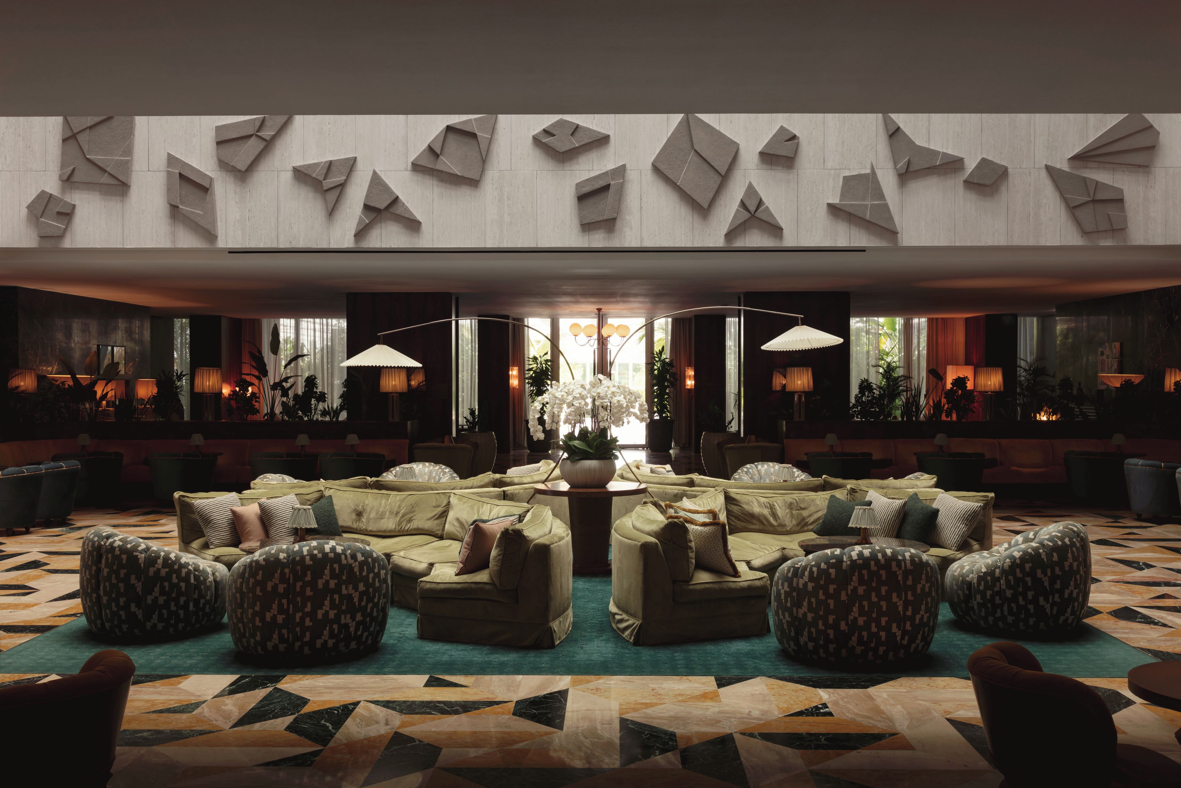 Een ruime lobby met een centraal zitgedeelte, grote ramen en een hoge plafond met geometrisch wanddecor.