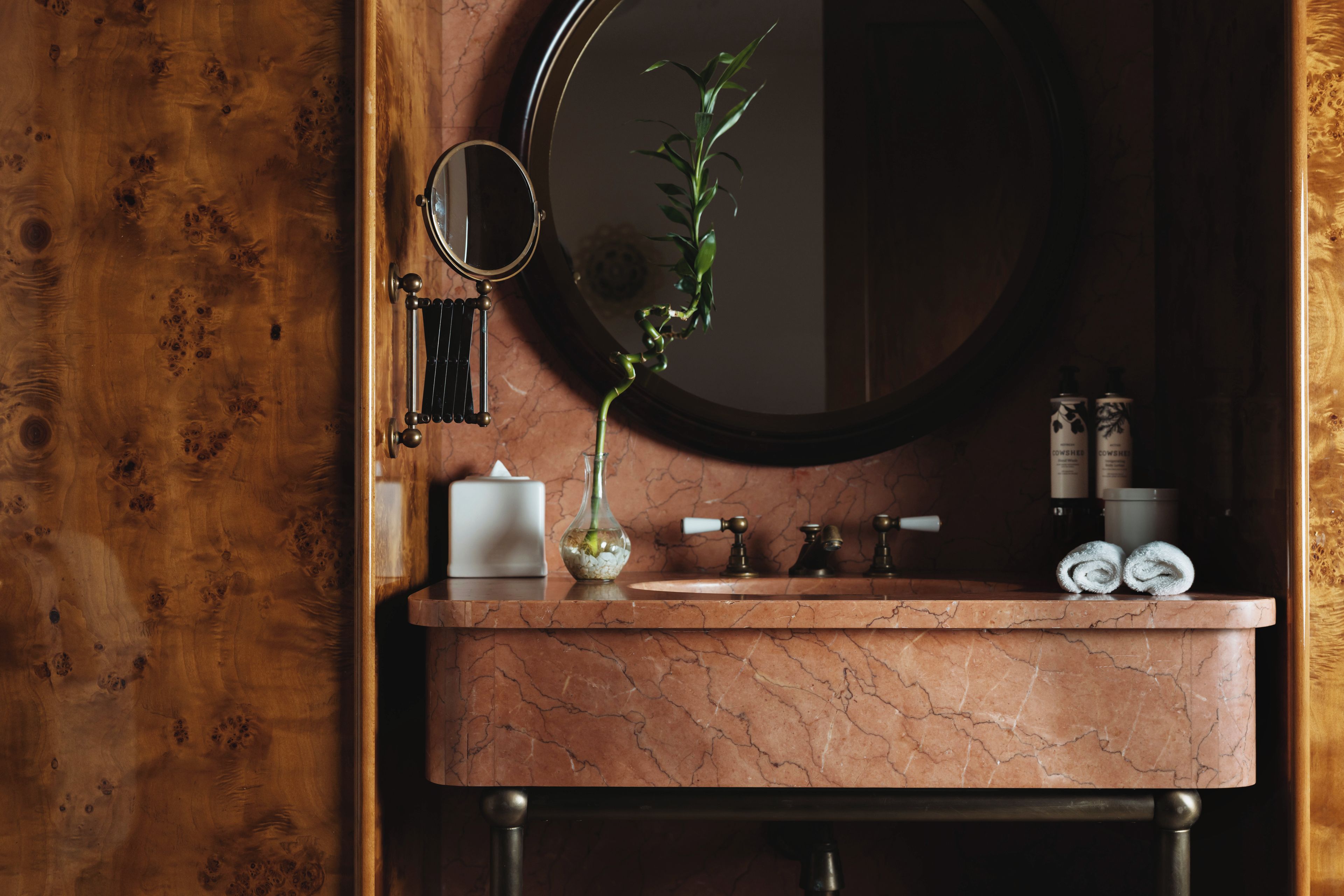 A bathroom with a wooden door, a round mirror, a marble sink, a vase with a plant, and toiletries.