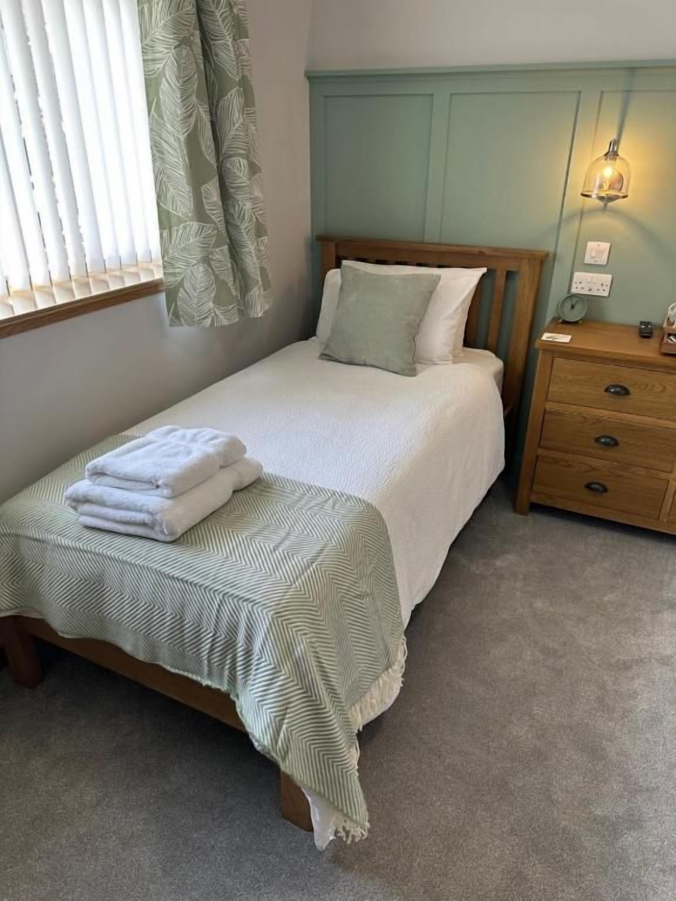 A single bed with a green and white striped bedspread, a wooden nightstand with drawers, a wall-mounted lamp, and a window with blinds.