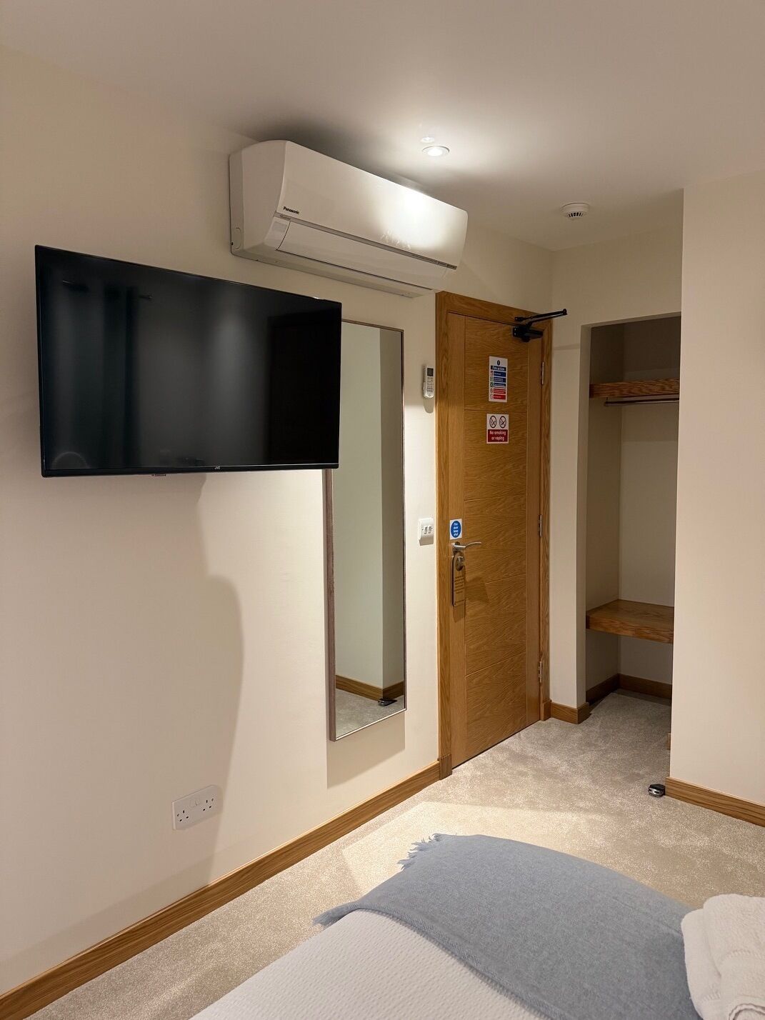 A hotel room with a flat-screen TV mounted on the wall, a wooden door with a mirror, and a closet with a shelf.