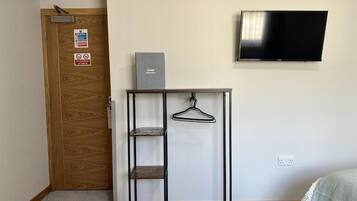 A room with a wooden door, a metal shelf unit with a hanger, a flat-screen TV, and a bed with a striped blanket.