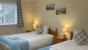 A room with two single beds, each with a blue bedspread and white pillows, a bedside table with a lamp, and a window with blinds and a curtain.