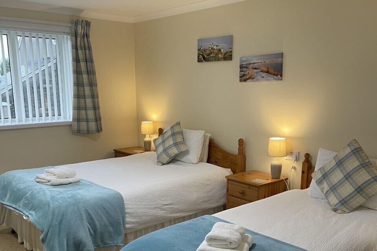 Ein Zimmer mit zwei Einzelbetten, jeweils mit blauer Bettdecke und weißen Kissen, einem Nachttisch mit Lampe sowie einem Fenster mit Jalousien und Vorhang.
