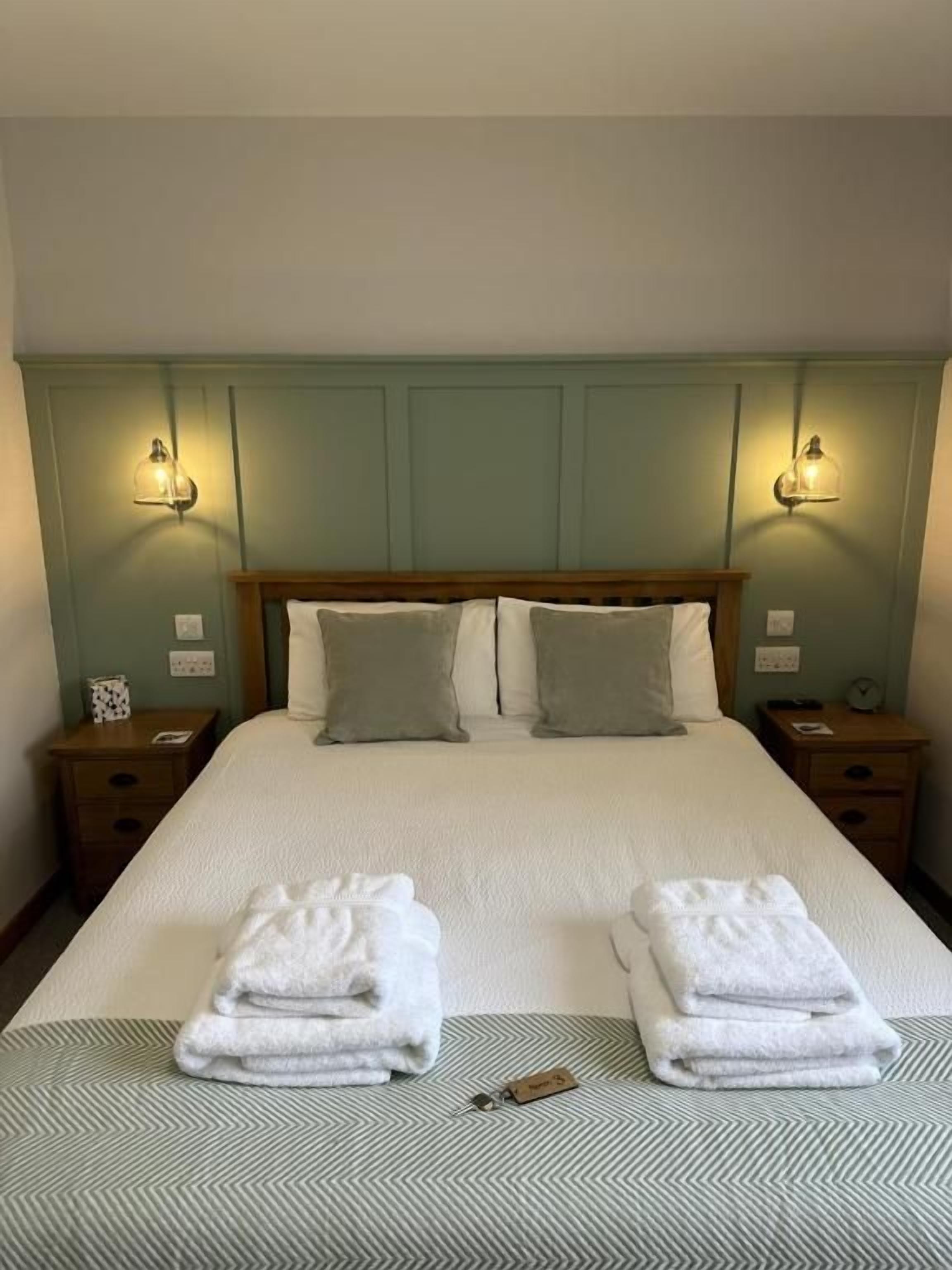 Un lit bien fait, avec deux oreillers, une serviette pliée et une clé posée sur le lit. Il y a deux tables de chevet, chacune avec une lampe, de chaque côté du lit.