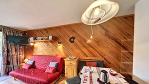Ein gemütliches Zimmer mit einem roten Sofa, einem Holzschrank, einem Esstisch mit Tischdecke und einem an der Wand befestigten Fernseher.