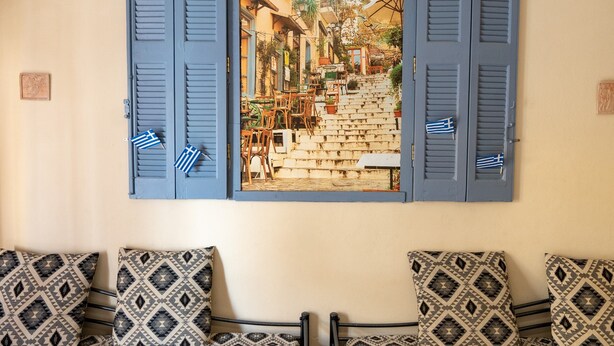 Une chambre avec un lit, des coussins à motifs et une vue sur une cour, visible à travers des volets bleus, où se trouvent des meubles d’extérieur et un escalier.
