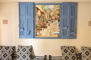Une chambre avec un lit, des coussins à motifs et une vue sur une cour, visible à travers des volets bleus, où se trouvent des meubles d’extérieur et un escalier.