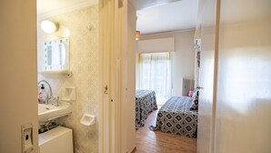Un couloir étroit mène à une chambre à coucher qui contient un lit, une table de chevet et une fenêtre avec des rideaux.
