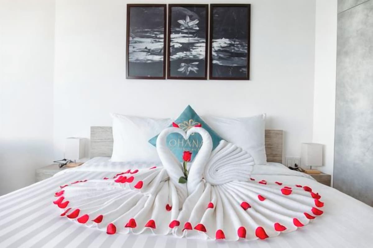 A bed with a swan-shaped pillow and a swan-shaped bedspread, a wall with three framed pictures, and a nightstand with a lamp.