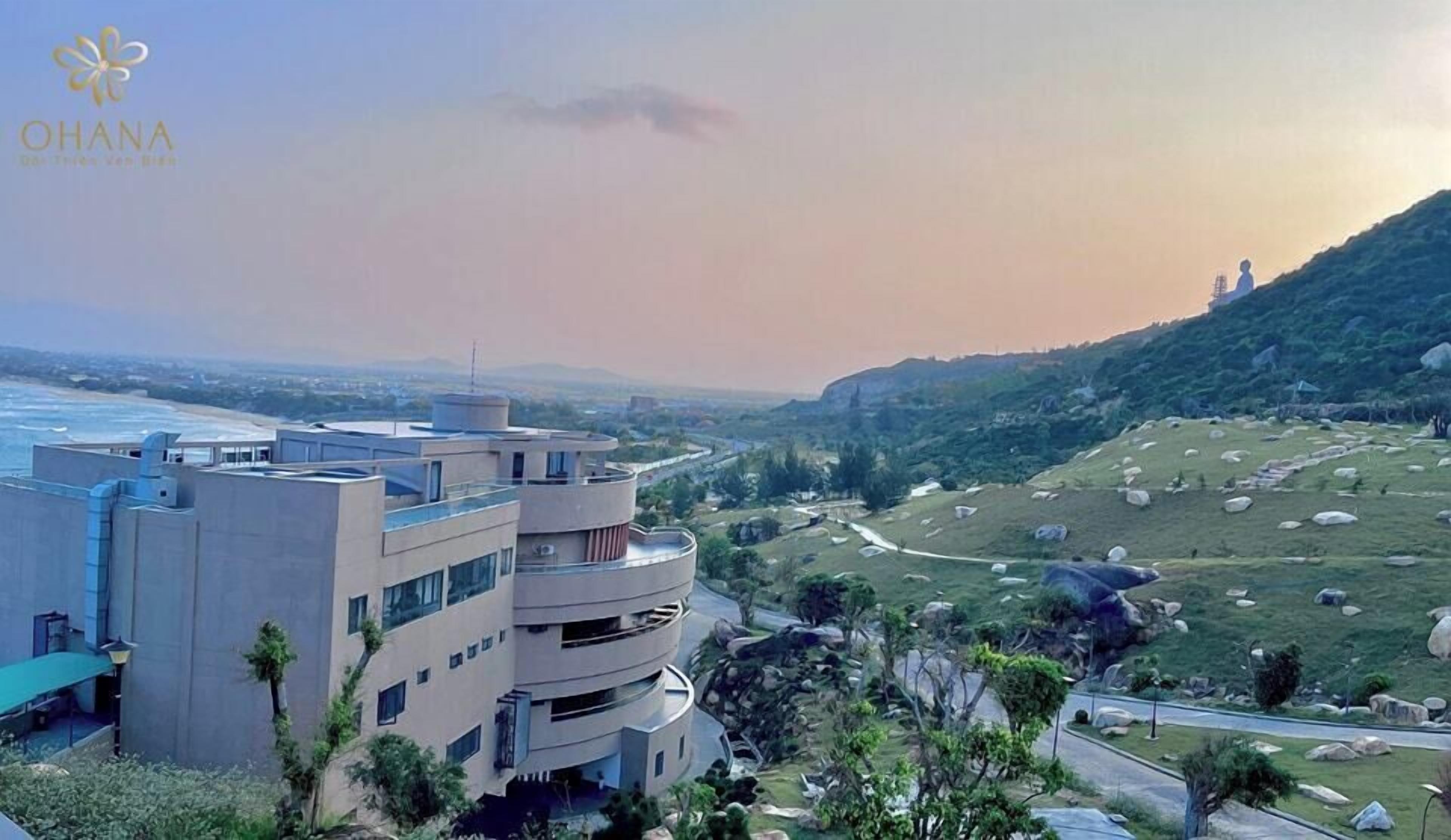 A modern building with multiple floors, a flat roof, and a view of the ocean and hills in the background.