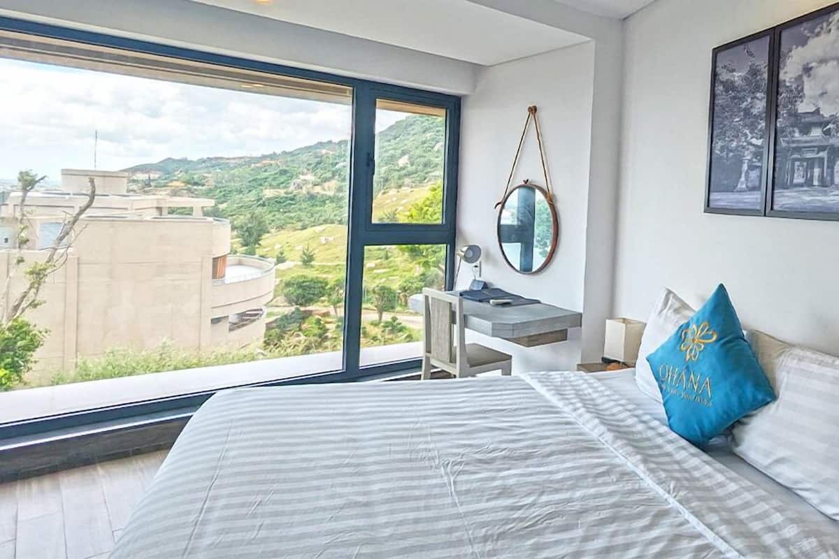 Une chambre avec une grande fenêtre, un lit avec une literie à rayures, un bureau avec une chaise et un miroir fixé au mur.