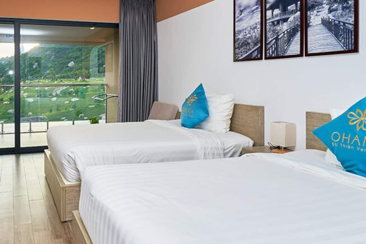 Une chambre d’hôtel avec deux lits, un bureau, une chaise, une télévision et un balcon donnant sur un espace verdoyant.