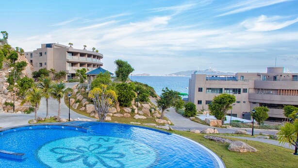 A large, circular swimming pool with a clear blue center, surrounded by a paved walkway and palm trees. - Ohana Village (Cat Tien)