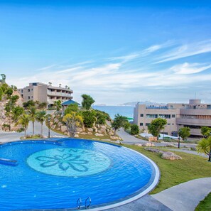 A large, circular swimming pool with a clear blue center, surrounded by a paved walkway and palm trees. - Ohana Village (Cat Tien)