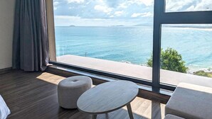 A room with a large window overlooking a body of water, a wooden floor, and a round table with two ottomans. - Ohana Village (Cat Tien)