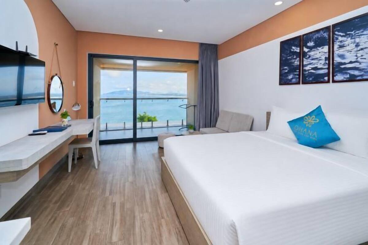 Habitación de hotel con cama, escritorio, silla, televisor y ventana grande con vista al mar.