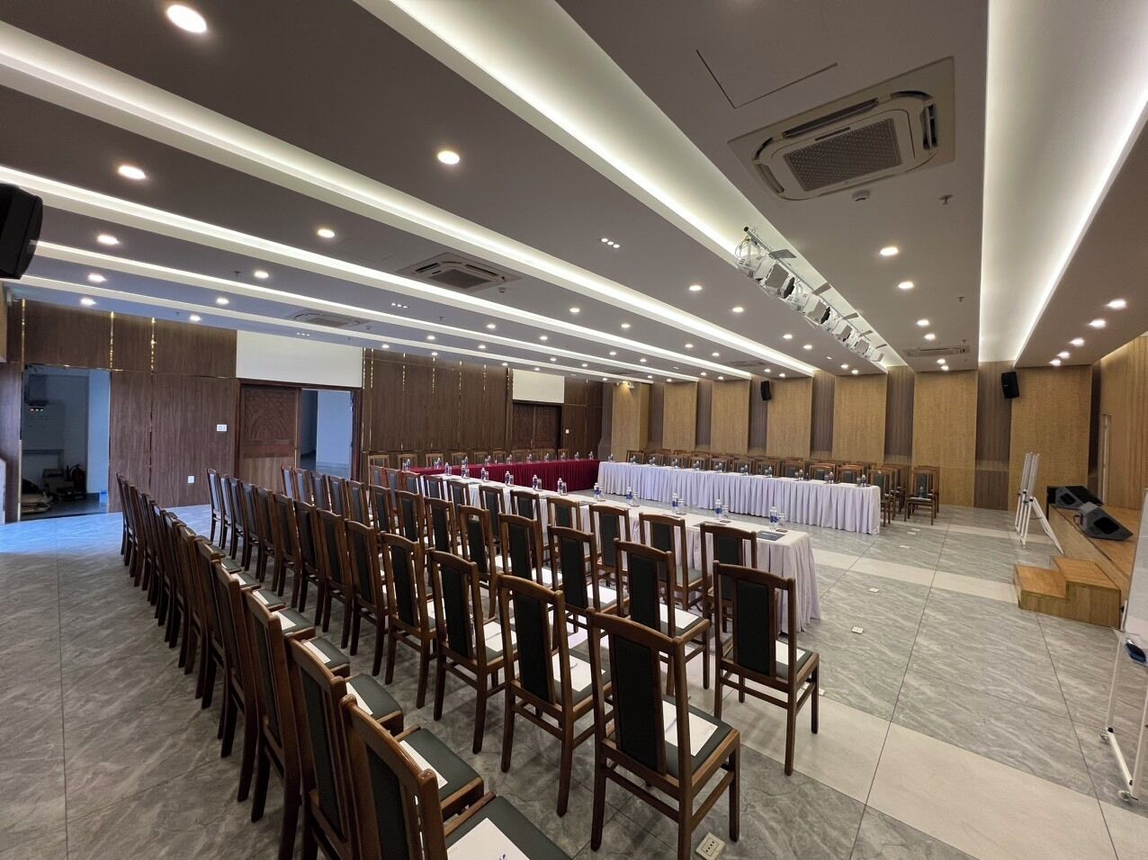 Une salle de conférence avec des rangées de chaises en bois, des tables recouvertes de nappes blanches et une scène dotée d’un pupitre.