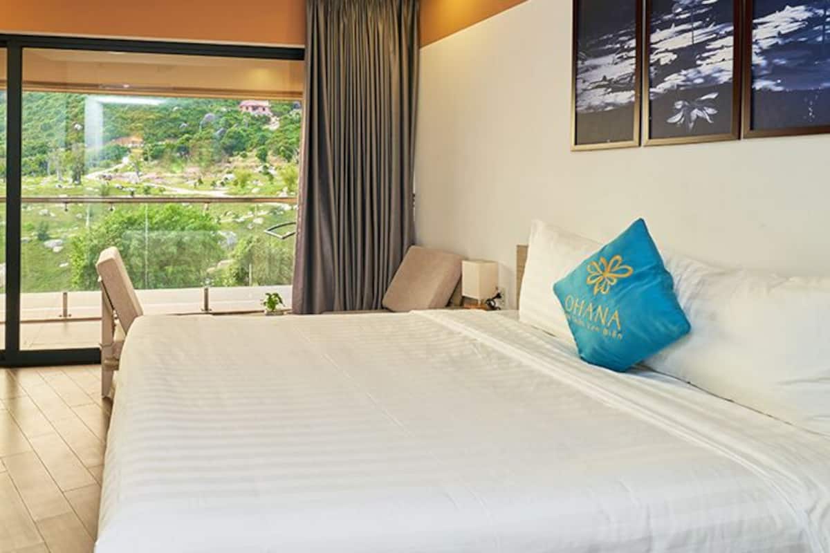 Une chambre d’hôtel avec un lit, un bureau et une vue sur un paysage luxuriant, visible à travers une grande fenêtre.