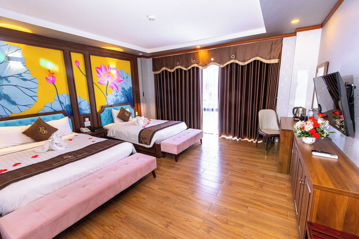 A hotel room with two beds, a wooden floor, a desk with a TV, and a large window with curtains.