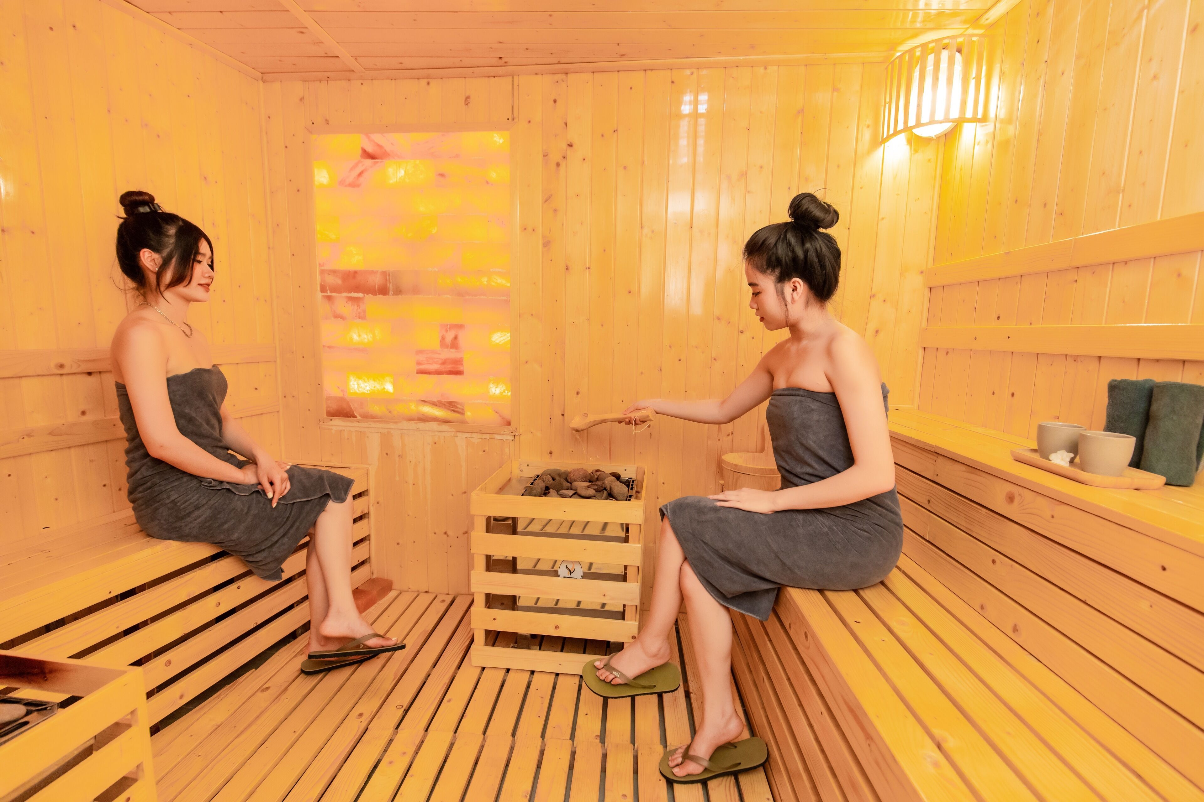 Two women in a wooden sauna room with benches, a wooden crate, and a wall-mounted heater.