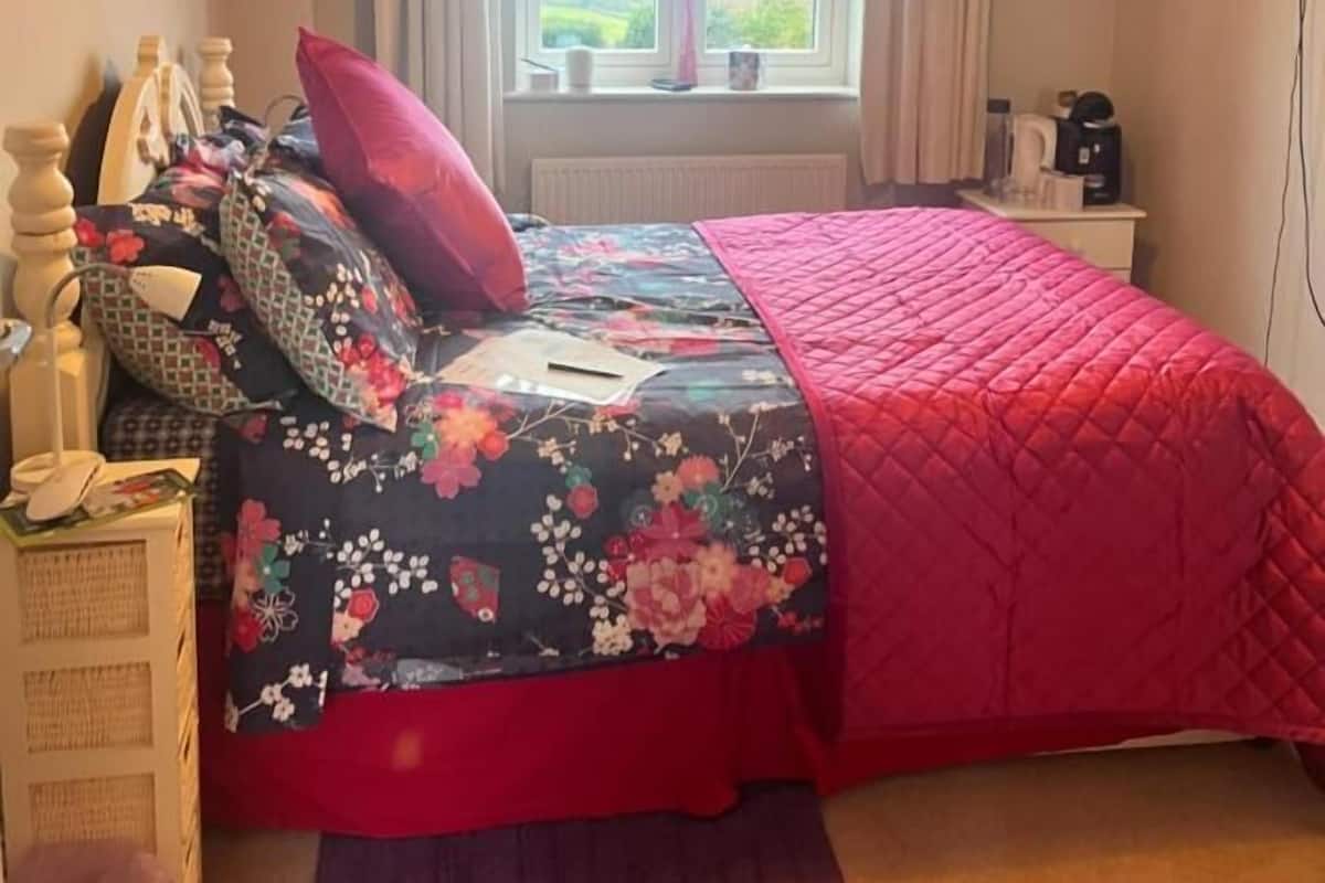 A bedroom with a bed, a window with curtains, a bedside table, a lamp, and a television on the wall.