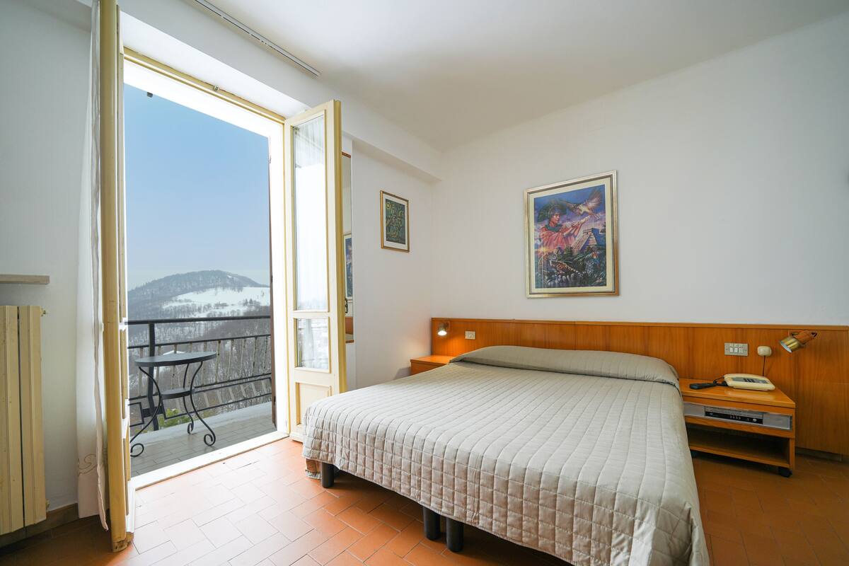 A bedroom with a bed, a nightstand, a lamp, a painting, and a view of a snowy landscape through an open door.