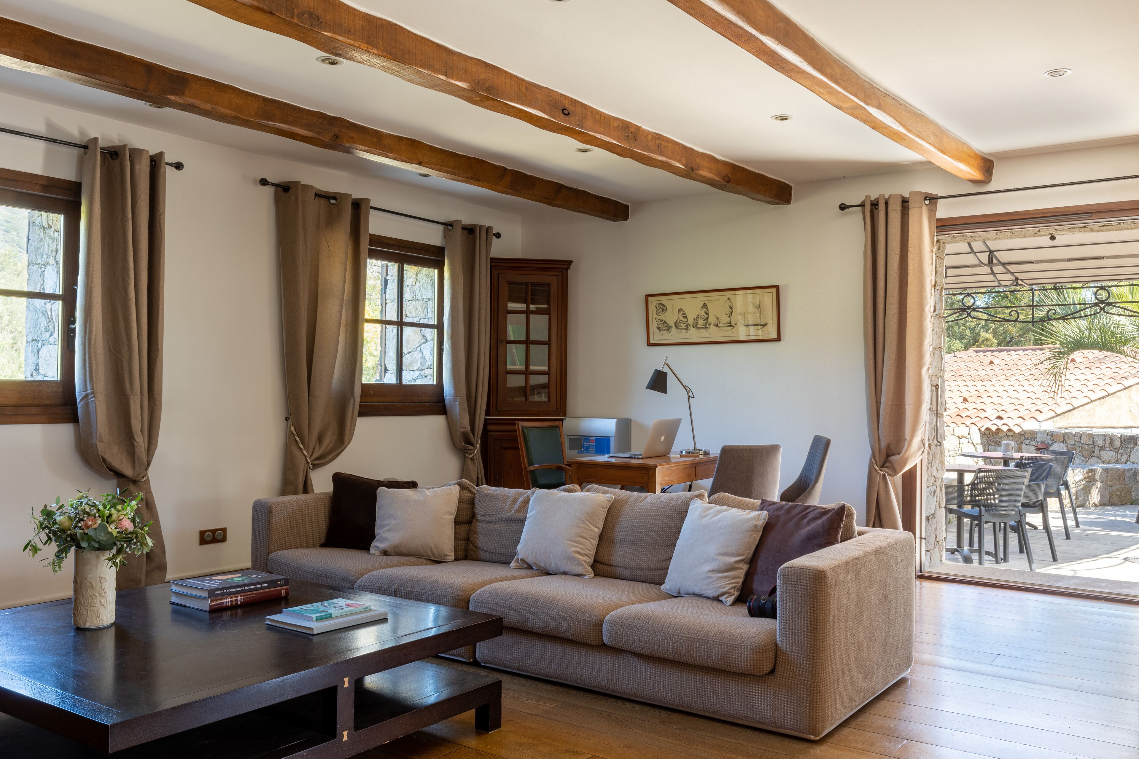 Un salon avec un canapé, une table basse et un bureau avec un ordinateur portable. Il y a une fenêtre avec des rideaux, un tableau encadré au mur et une vue sur l’extérieur.