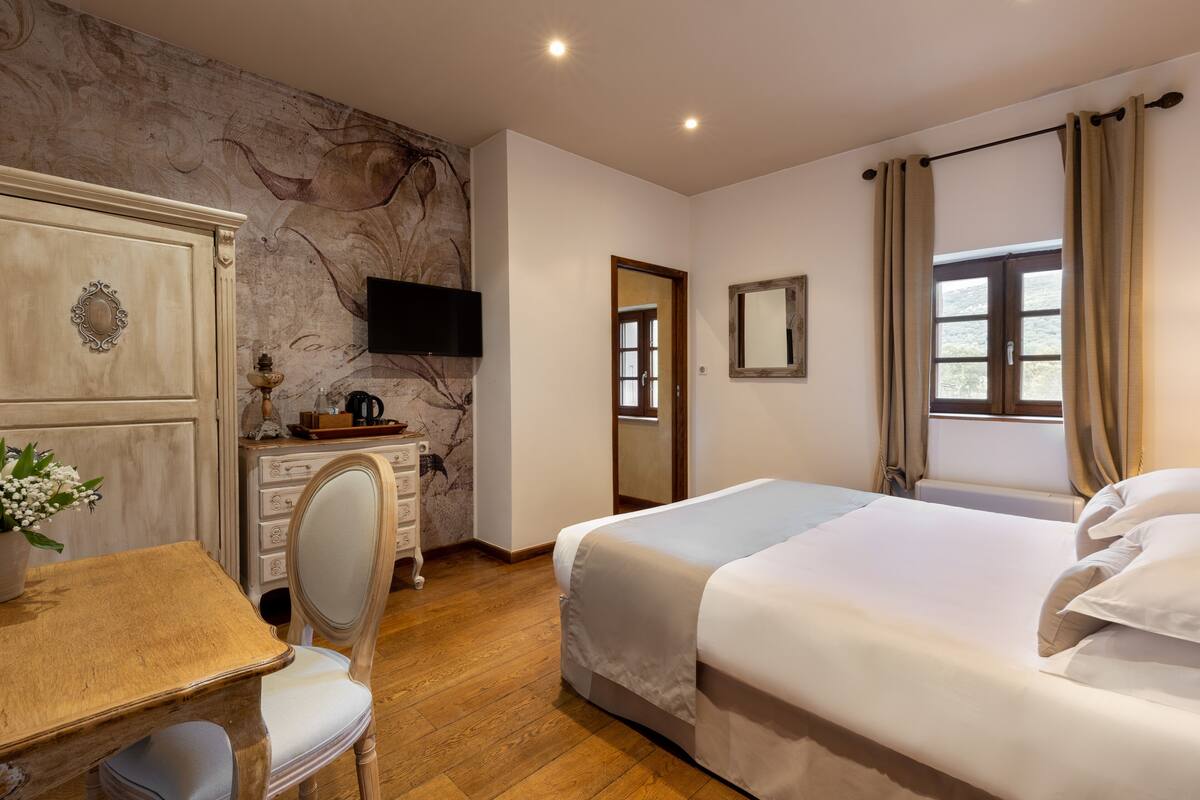 Ein Hotelzimmer mit einem Holzkasten, einem Schreibtisch mit Stuhl, einem Bett mit weißer Bettwäsche, einem an der Wand befestigten Flachbildfernseher und einem Fenster mit Vorhängen.