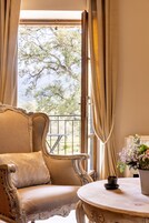 Quarto com uma cadeira clássica, uma mesa com um vaso de flores e vista para árvores através de uma janela.