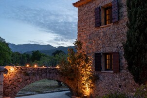 Um edifício de pedra com uma ponte de pedra, rodeado por vegetação e montanhas ao fundo.