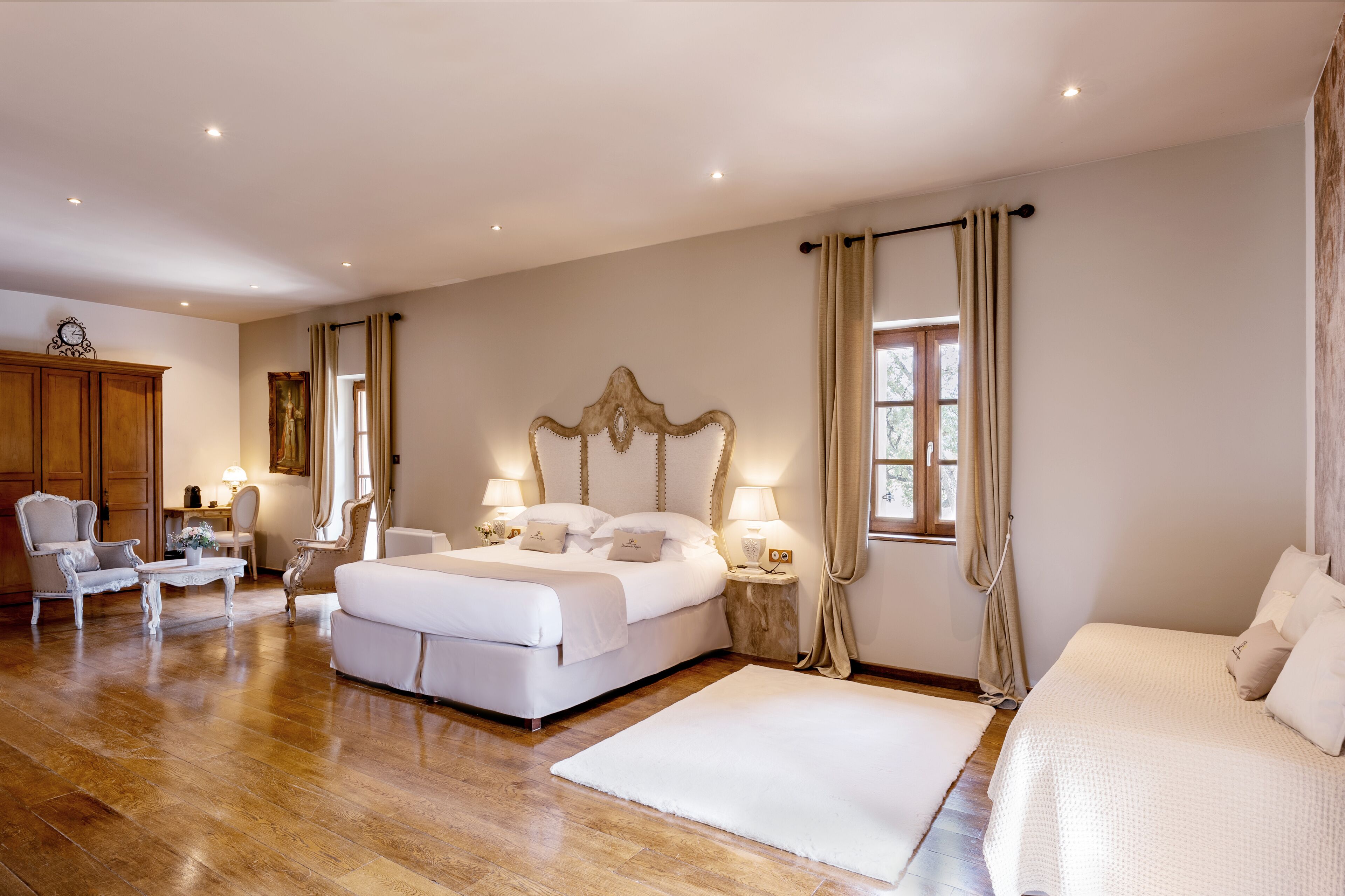 A spacious bedroom with a large bed, two bedside tables with lamps, a wooden wardrobe, and a seating area with a chair and table.