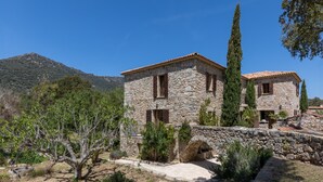 Edifici de pedra amb teulada a falda, envoltat d'arbres i una muntanya al fons.