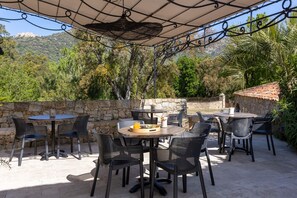 An outdoor patio with round tables and chairs, surrounded by stone walls and trees. - Domaine du Reginu (Feliceto)