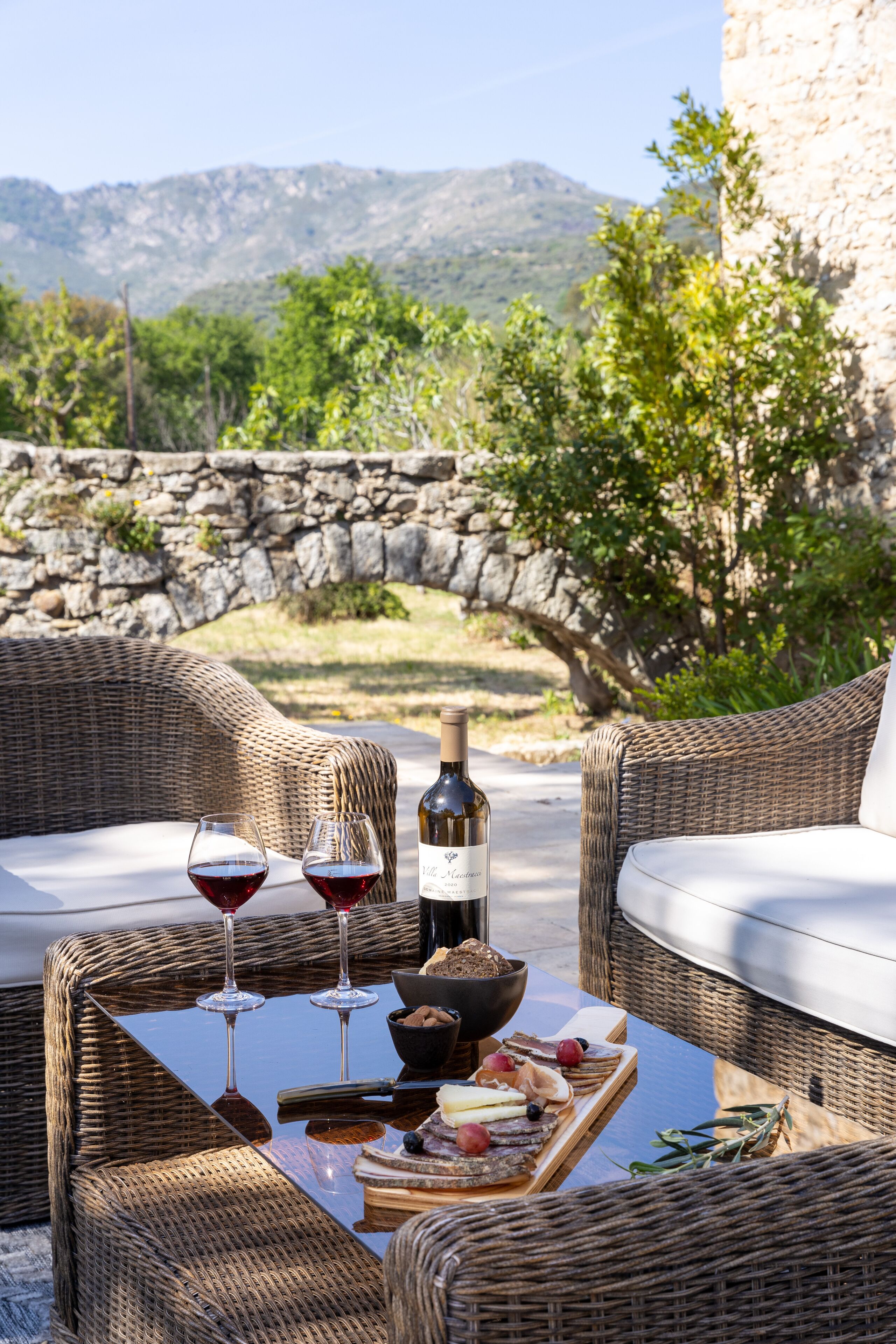 A wicker table set with wine glasses, a bottle, and a cheese board.