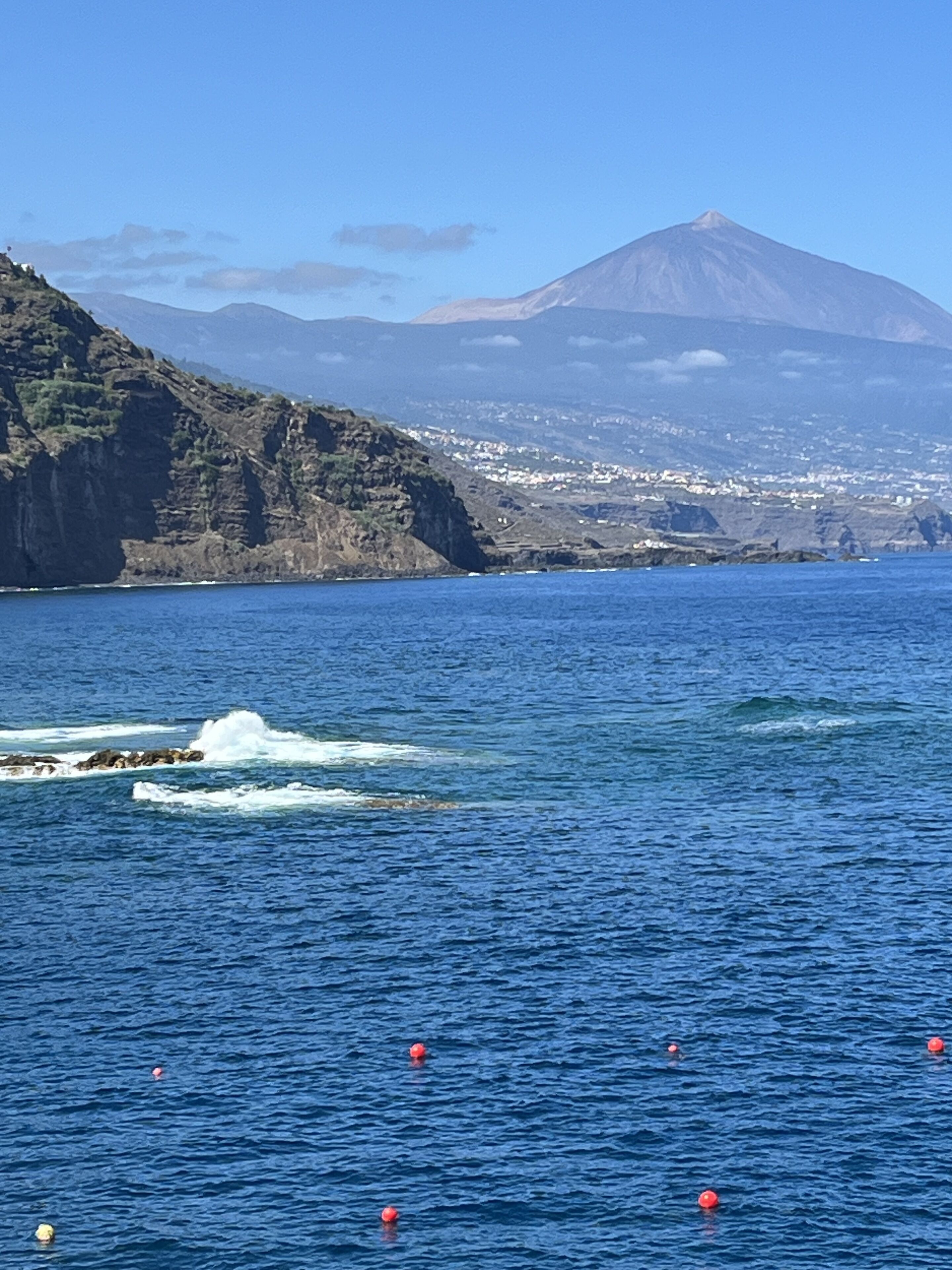 A coastal landscape with a mountain in the background, clear blue water, and red buoys.