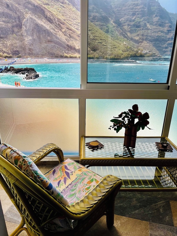 Une pièce dotée d’une grande fenêtre donnant sur une zone côtière avec une plage et des falaises rocheuses. À l’intérieur, on trouve un fauteuil en osier agrémenté d’un coussin coloré et une petite table sur laquelle est posé un vase de fleurs.